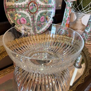 May include: A clear, cut-glass bowl with a starburst design, resting on a mirrored tray. A decorative plate with pink floral and bird motifs is in the background, along with a candle holder and a decorative wire basket.