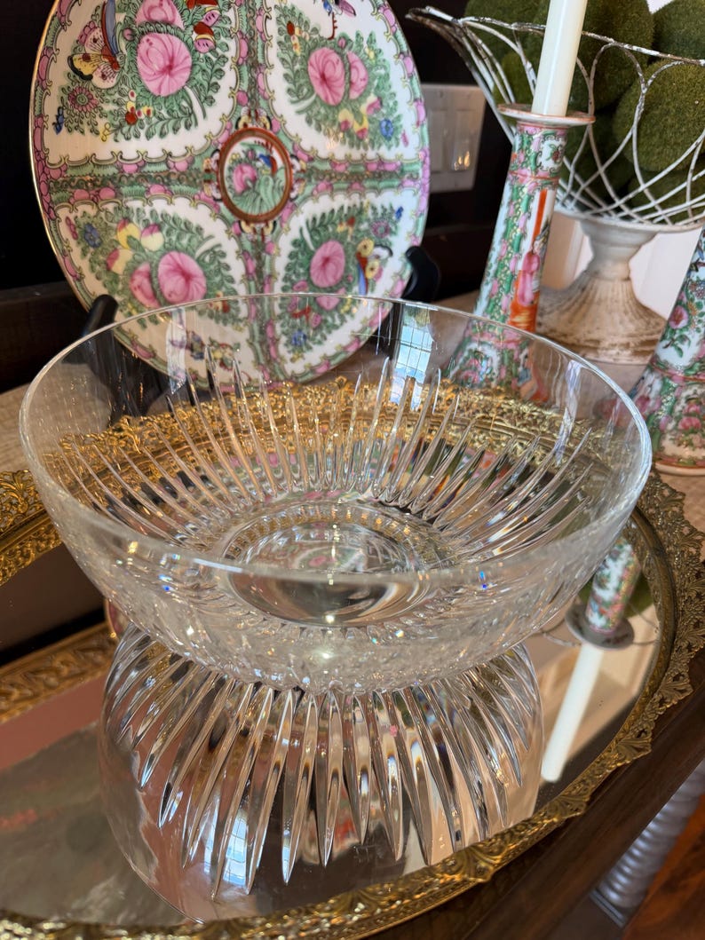 May include: A clear, cut-glass bowl with a starburst design, resting on a mirrored tray. A decorative plate with pink floral and bird motifs is in the background, along with a candle holder and a decorative wire basket.