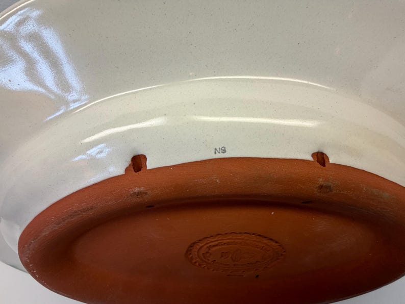Puede incluir: La parte inferior de un cuenco de cer&aacute;mica con una superficie interior lisa esmaltada en color blanco roto y una base de color terracota sin esmaltar. La base incluye dos peque&ntilde;as aberturas rectangulares y el texto N9 en color oscuro.