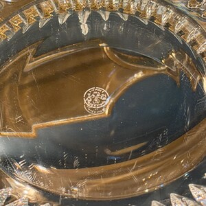 May include: Close-up of a clear crystal glass object with intricate, geometric patterns. The rim features a series of pointed, leaf-like shapes. The base has a circular emblem with text. The glass reflects light, creating a sparkling effect.