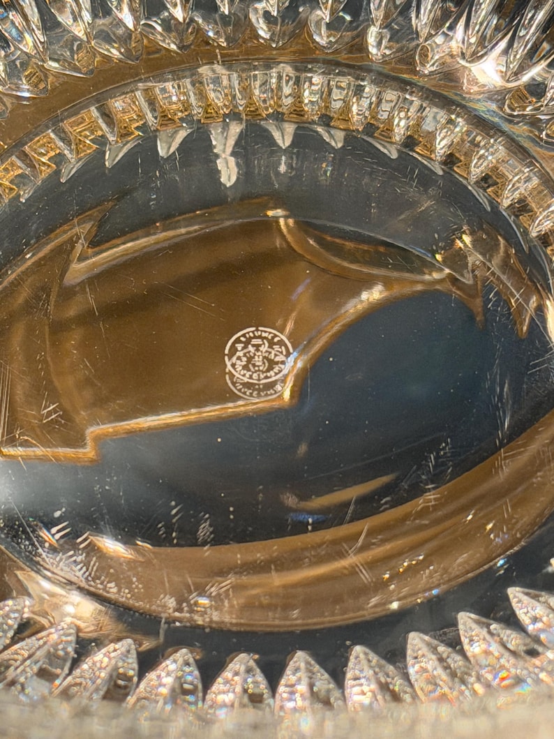 May include: Close-up of a clear crystal glass object with intricate, geometric patterns. The rim features a series of pointed, leaf-like shapes. The base has a circular emblem with text. The glass reflects light, creating a sparkling effect.