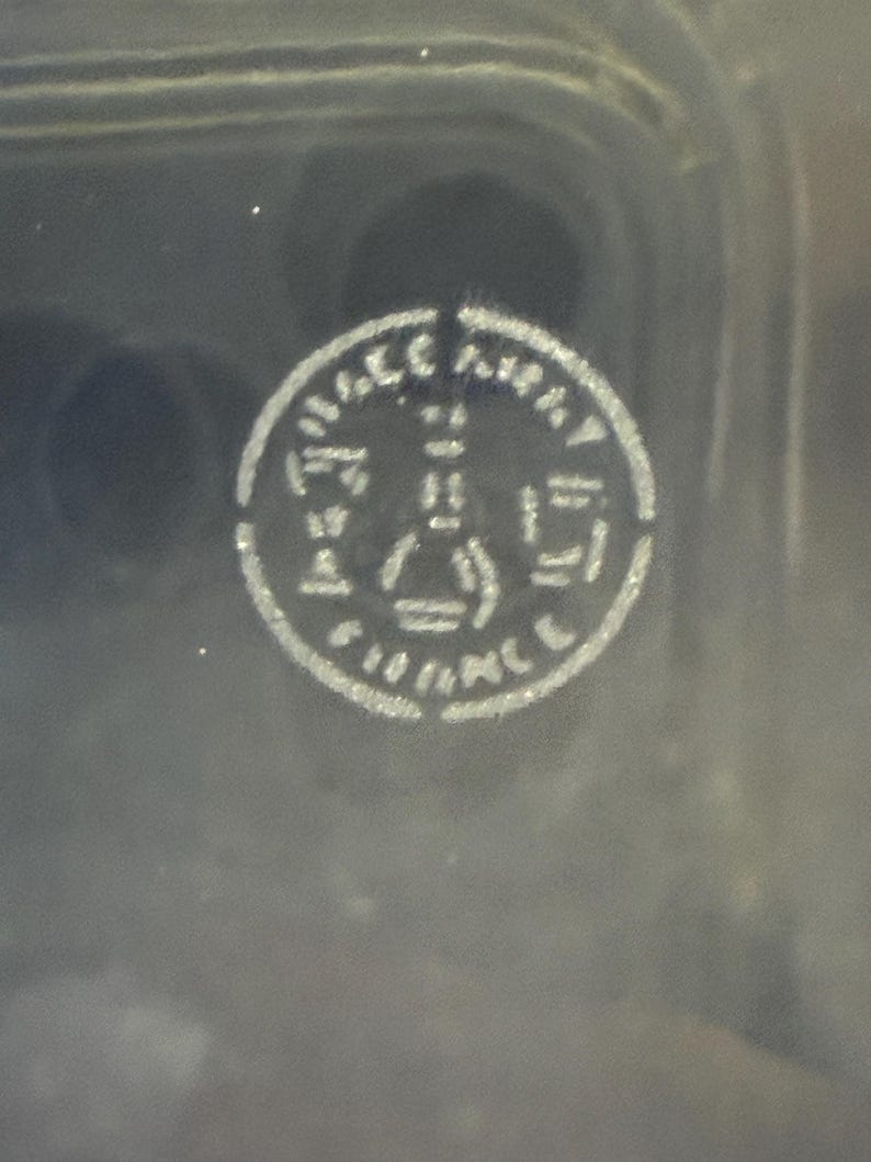 May include: A circular, white emblem with the text "FRANCE" and other indiscernible text. The emblem features a central design with geometric shapes, including a triangle and a rectangle. The background is a dark, possibly black, surface.