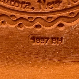 Puede incluir: Una superficie de cer&aacute;mica color terracota con un sello en relieve. El texto dice Aurora, New York, con la fecha 1997 BH estampada debajo. Una l&iacute;nea ondulada decorativa con muescas circulares separa el texto de la fecha estampada.