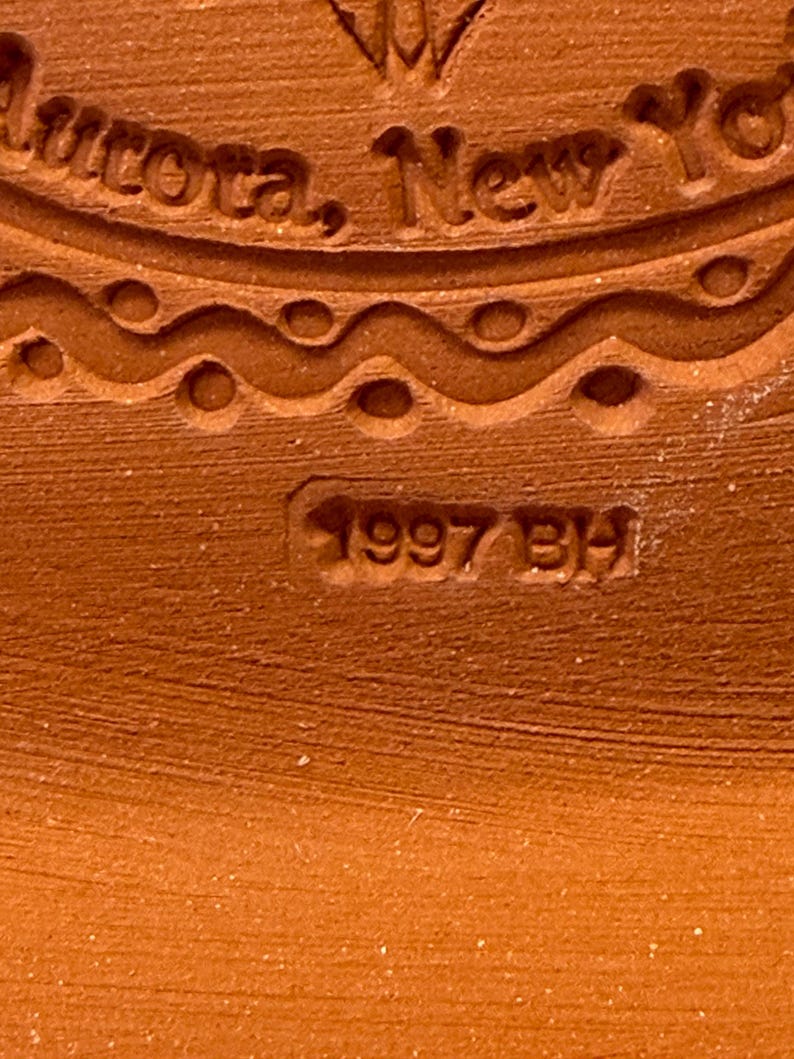 Puede incluir: Una superficie de cer&aacute;mica color terracota con un sello en relieve. El texto dice Aurora, New York, con la fecha 1997 BH estampada debajo. Una l&iacute;nea ondulada decorativa con muescas circulares separa el texto de la fecha estampada.