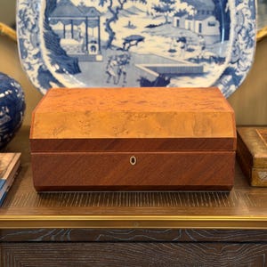 Może przedstawiać: Prostokątne drewniane pudełko z dwutonowym wykończeniem. Górna część wykonana jest z jaśniejszego, wzorzystego drewna, a dolna z ciemniejszego, litego drewna. Mała, okrągła dziurka od klucza w kolorze złotym jest wyśrodkowana z przodu. Pudełko jest prezentowane przed niebiesko-białym talerzem dekoracyjnym.