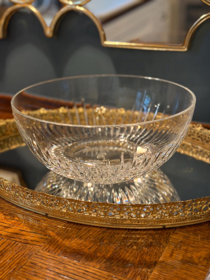 May include: A clear glass bowl with a textured, ribbed design. The bowl sits on a gold-colored, ornate tray. The bowl is reflecting the light, creating a sparkling effect. The bowl is likely used for serving or display.
