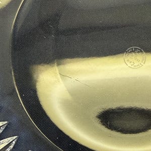 May include: Close-up of a clear glass plate with a decorative starburst design on the rim. The plate has a dark, reflective surface and a small, circular stamp near the center.