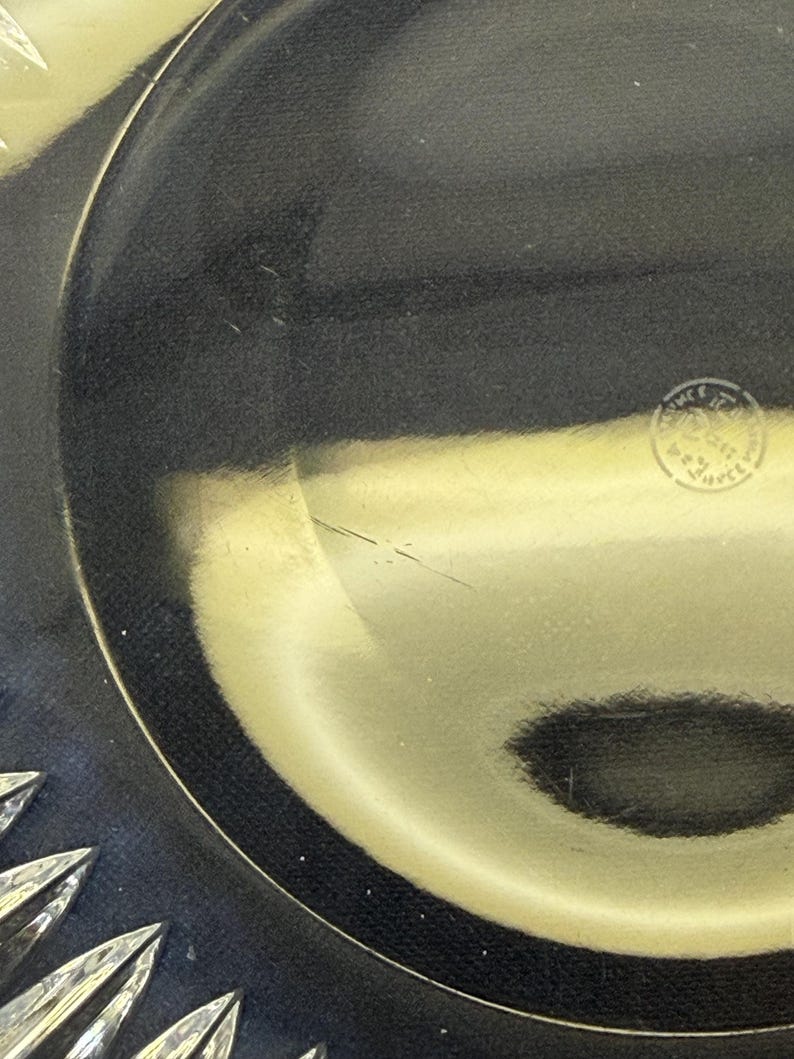 May include: Close-up of a clear glass plate with a decorative starburst design on the rim. The plate has a dark, reflective surface and a small, circular stamp near the center.