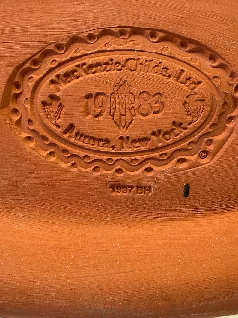 Puede incluir: Una base de cer&aacute;mica de color terracota con un sello ovalado en relieve que dice MacKenzie-Childs, Ltd, 1983, Aurora, New York, con un logotipo central y espigas de trigo. Debajo del &oacute;valo, el texto 1997 BH est&aacute; grabado en la arcilla.