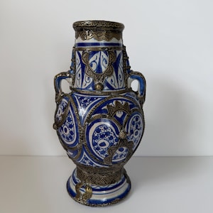 May include: A decorative ceramic vase with a blue and white floral pattern. The vase has two handles and is accented with silver-colored metal trim. The vase is approximately 10 inches tall.
