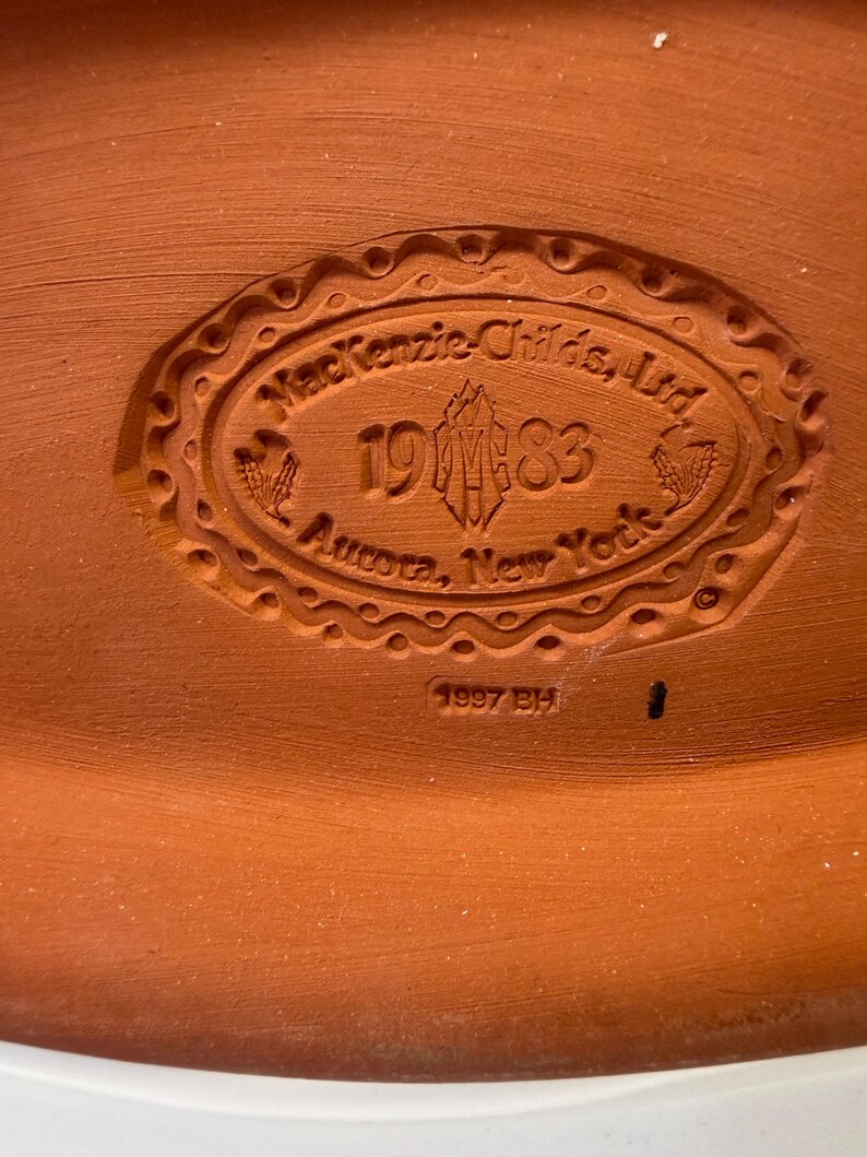 Puede incluir: Una base de cer&aacute;mica color terracota con un sello ovalado en relieve que dice MacKenzie-Childs, Ltd. 1983 Aurora, New York, con un logotipo central y motivos de trigo. Debajo del &oacute;valo, el texto 1997 BH est&aacute; claramente impreso en la arcilla.