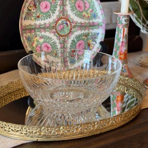 May include: A clear glass bowl with a textured design sits on a gold-rimmed mirror tray. Behind it, a decorative plate with pink floral and bird patterns is displayed. A candle holder with a matching design is also visible.