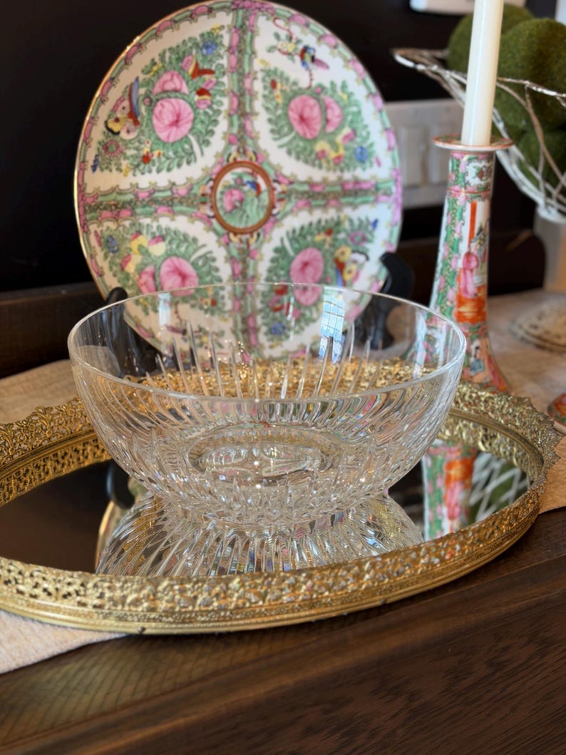 May include: A clear glass bowl with a textured design sits on a gold-rimmed mirror tray. Behind it, a decorative plate with pink floral and bird patterns is displayed. A candle holder with a matching design is also visible.