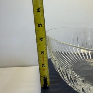 May include: A clear glass bowl with a textured, patterned design. The bowl is approximately 6 inches tall, as measured by a yellow measuring tape. The bowl is on a dark surface.