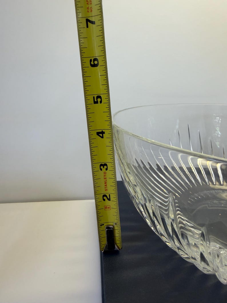 May include: A clear glass bowl with a textured, patterned design. The bowl is approximately 6 inches tall, as measured by a yellow measuring tape. The bowl is on a dark surface.