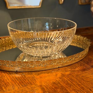 May include: A clear glass bowl with a textured, ribbed design sits on a gold-colored, ornate, mirrored tray. The bowl reflects the light, and the tray has a decorative, filigree edge. The background includes a gold-framed mirror.