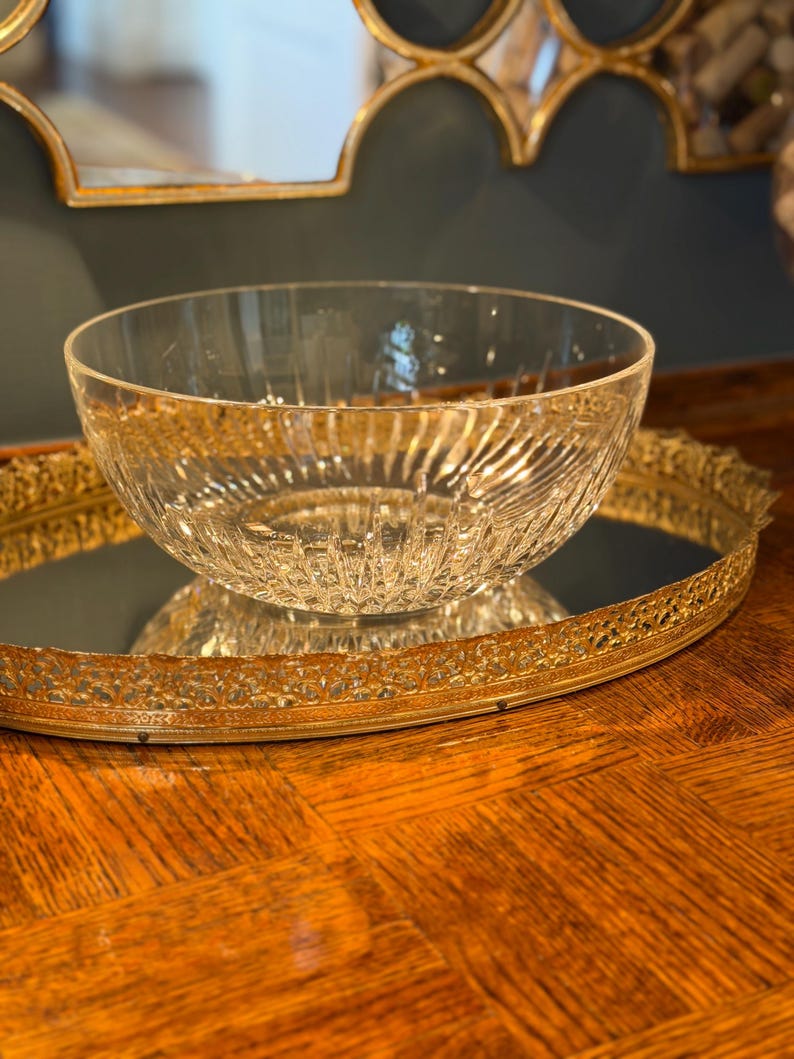 May include: A clear glass bowl with a textured, ribbed design sits on a gold-colored, ornate, mirrored tray. The bowl reflects the light, and the tray has a decorative, filigree edge. The background includes a gold-framed mirror.