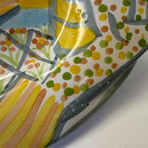 Puede incluir: Un cuenco de cer&aacute;mica con un dise&ntilde;o pintado a mano que presenta formas abstractas en amarillo, naranja y verde, incluyendo patrones de rayas y grupos de peque&ntilde;os puntos sobre un fondo blanco, con contornos gris oscuro definiendo los motivos.