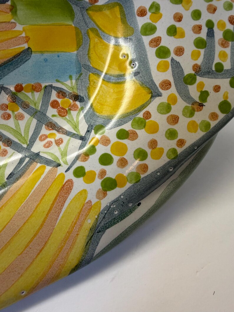 Puede incluir: Un cuenco de cer&aacute;mica con un dise&ntilde;o pintado a mano que presenta formas abstractas en amarillo, naranja y verde, incluyendo patrones de rayas y grupos de peque&ntilde;os puntos sobre un fondo blanco, con contornos gris oscuro definiendo los motivos.