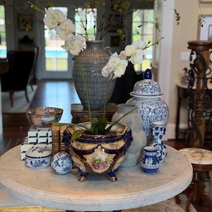 Puede incluir: Una colección de objetos decorativos sobre una mesa redonda, que incluye una maceta de cerámica azul y dorada con motivos florales, un tarro de jengibre azul y blanco, un jarrón gris texturizado, un cuenco pequeño y una orquídea blanca.