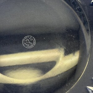 May include: Close-up of a clear glass plate with a decorative starburst pattern around the edge. The plate has a circular shape and a smooth surface. A small, circular emblem is visible in the center.