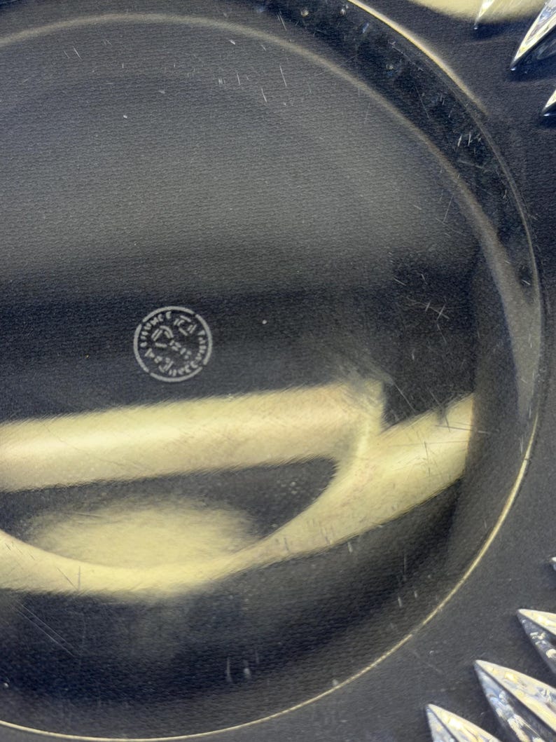 May include: Close-up of a clear glass plate with a decorative starburst pattern around the edge. The plate has a circular shape and a smooth surface. A small, circular emblem is visible in the center.
