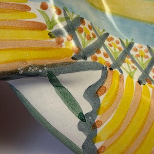 Puede incluir: Un cuenco de cer&aacute;mica con un dise&ntilde;o pintado a mano de formas amarillas similares a pl&aacute;tanos, bayas naranjas y follaje verde. La superficie tiene un acabado brillante con una base blanca, acentuada por l&iacute;neas onduladas de color gris oscuro.