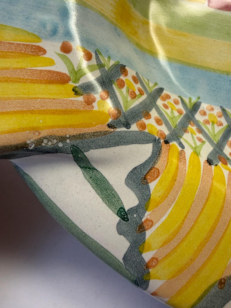 Puede incluir: Un cuenco de cer&aacute;mica con un dise&ntilde;o pintado a mano de formas amarillas similares a pl&aacute;tanos, bayas naranjas y follaje verde. La superficie tiene un acabado brillante con una base blanca, acentuada por l&iacute;neas onduladas de color gris oscuro.