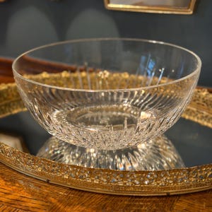 May include: A clear, decorative glass bowl with a textured, ridged design. The bowl sits on a gold-colored, ornate tray, reflecting its image. The background includes a dark wall and a gold-framed mirror.
