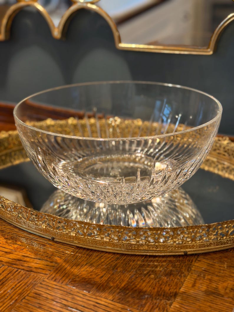 May include: A clear, decorative glass bowl with a textured, ridged design. The bowl sits on a gold-colored, ornate tray, reflecting its image. The background includes a dark wall and a gold-framed mirror.