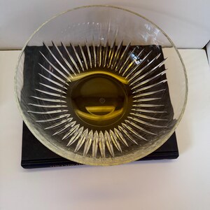 May include: A clear glass bowl with a sunburst design radiating from the center. The bowl has a wide, shallow shape and sits on a dark surface. The sunburst pattern is etched into the glass, creating a textured effect.