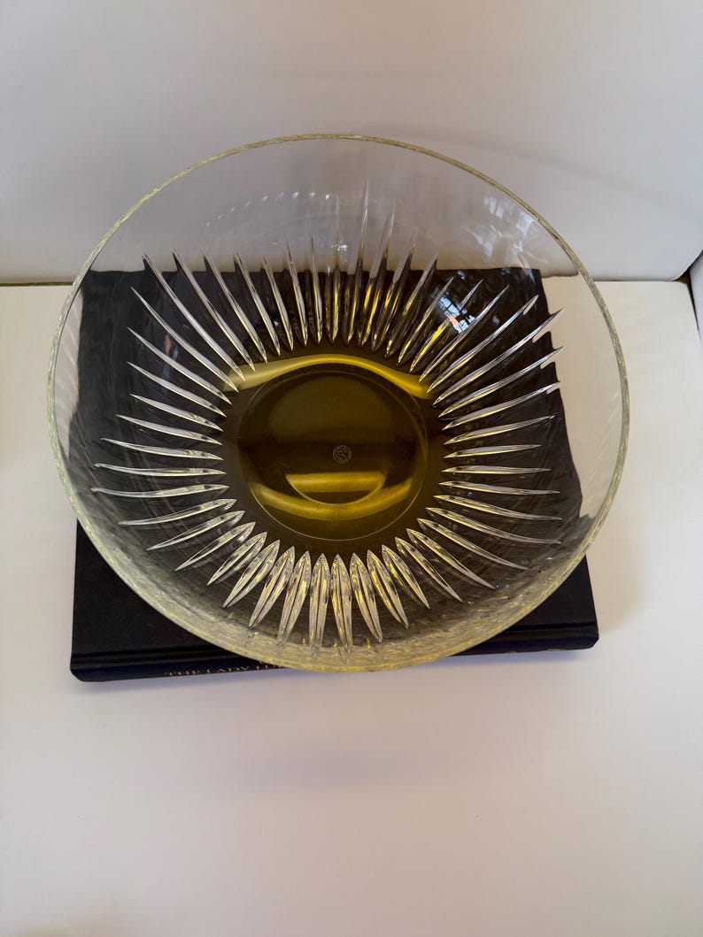May include: A clear glass bowl with a sunburst design radiating from the center. The bowl has a wide, shallow shape and sits on a dark surface. The sunburst pattern is etched into the glass, creating a textured effect.