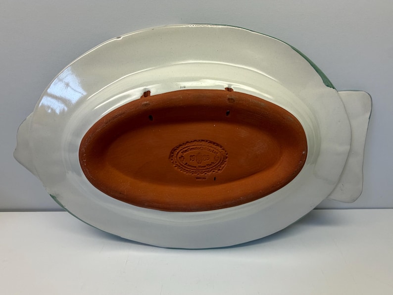Puede incluir: La parte inferior de una bandeja de cer&aacute;mica con forma de pez, con un acabado esmaltado blanco y una base central ovalada de terracota sin esmaltar. La secci&oacute;n de terracota tiene una marca de fabricante estampada y dos orificios para colgar.