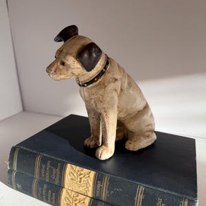 Puede incluir: Una figura de perro de hierro fundido con cuerpo color crema y orejas marrón oscuro, que lleva un collar negro con tachuelas plateadas. El perro está colocado sobre dos libros apilados de color azul oscuro con letras doradas.