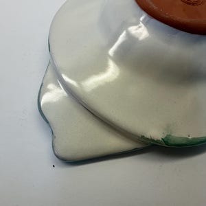 Puede incluir: Un plato de cer&aacute;mica con esmalte blanco brillante y un borde pintado en verde. La parte inferior presenta una base de terracota sin esmaltar con un logotipo circular estampado. El plato tiene forma de cuenco ancho y poco profundo con un asa plana.