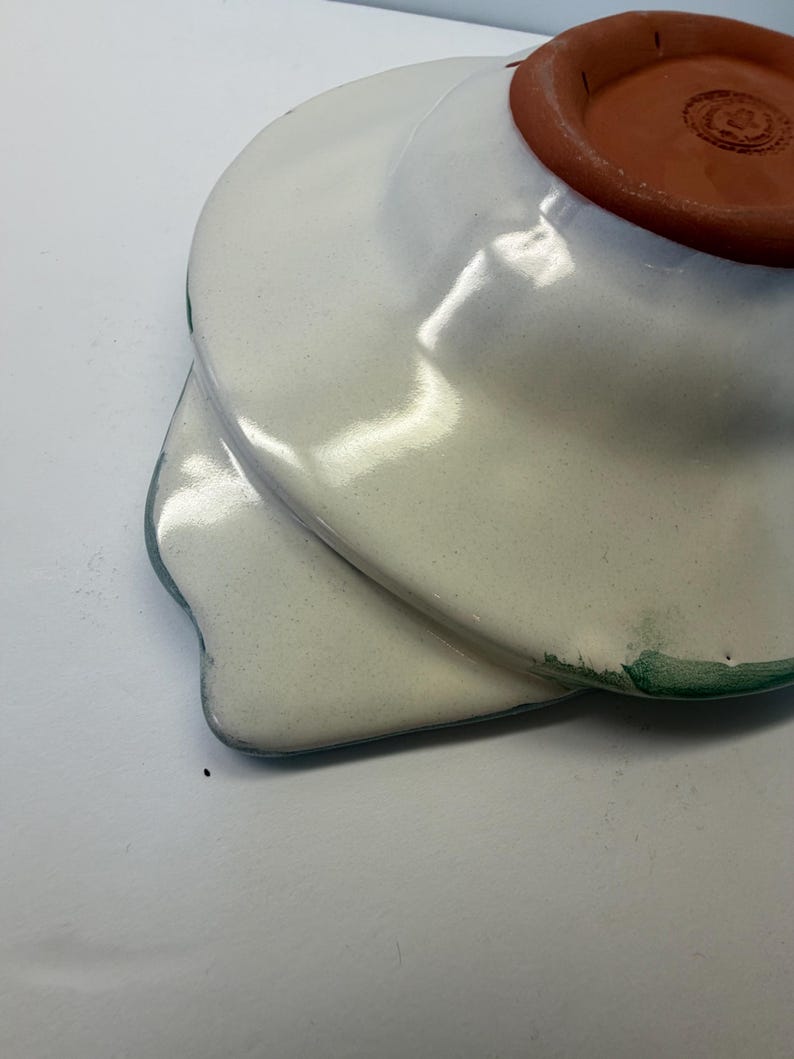 Puede incluir: Un plato de cer&aacute;mica con esmalte blanco brillante y un borde pintado en verde. La parte inferior presenta una base de terracota sin esmaltar con un logotipo circular estampado. El plato tiene forma de cuenco ancho y poco profundo con un asa plana.