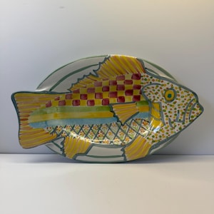 Puede incluir: Una fuente de servir ovalada de cer&aacute;mica con forma de pez, que presenta un dise&ntilde;o vibrante pintado a mano. El cuerpo muestra un patr&oacute;n de tablero de ajedrez rojo y amarillo, rayas horizontales en azul y verde, y un motivo floral amarillo en la parte inferior.