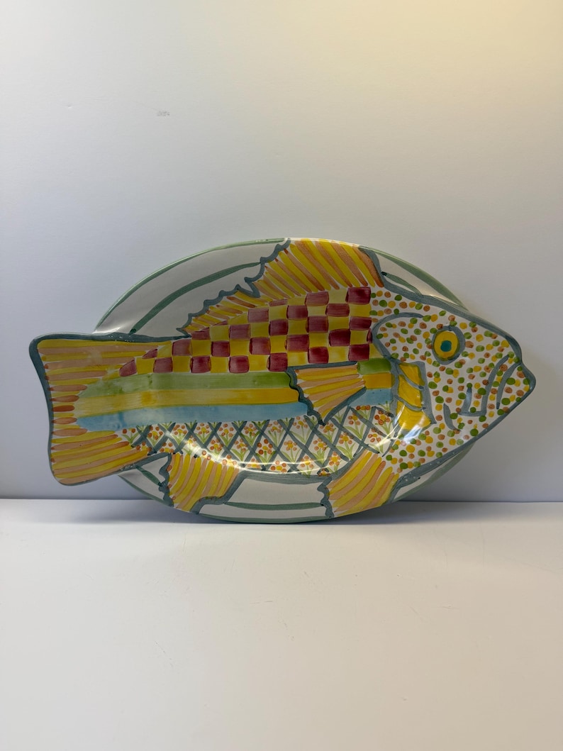 Puede incluir: Una fuente de servir ovalada de cer&aacute;mica con forma de pez, que presenta un dise&ntilde;o vibrante pintado a mano. El cuerpo muestra un patr&oacute;n de tablero de ajedrez rojo y amarillo, rayas horizontales en azul y verde, y un motivo floral amarillo en la parte inferior.