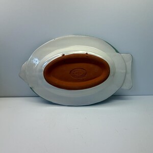 Puede incluir: La parte inferior de una bandeja de cer&aacute;mica con forma de pez, con un acabado esmaltado blanco y una base ovalada central de terracota sin esmaltar. La secci&oacute;n de terracota incluye una marca de fabricante y dos orificios para colgarla.