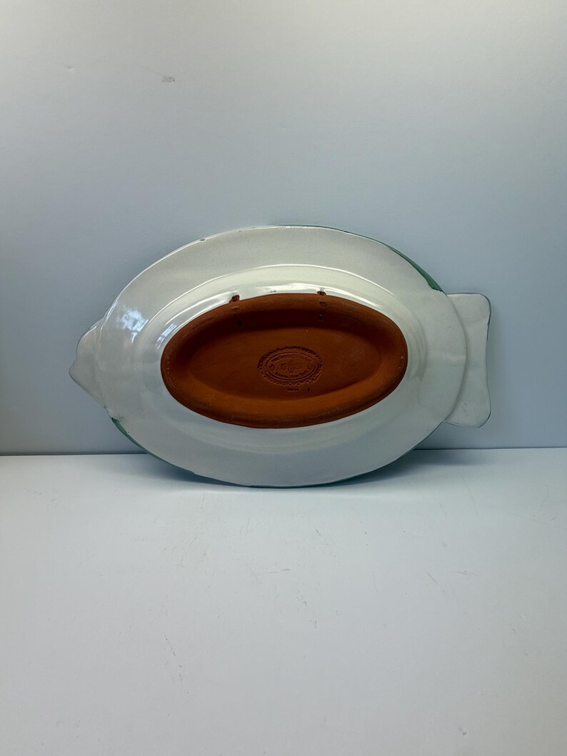 Puede incluir: La parte inferior de una bandeja de cer&aacute;mica con forma de pez, con un acabado esmaltado blanco y una base ovalada central de terracota sin esmaltar. La secci&oacute;n de terracota incluye una marca de fabricante y dos orificios para colgarla.