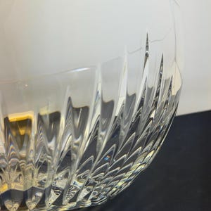 May include: A clear glass bowl with a textured, patterned design. The bowl's exterior features a series of angled, vertical cuts that create a repeating pattern. The bowl is partially lit, with light reflecting off the textured surface.