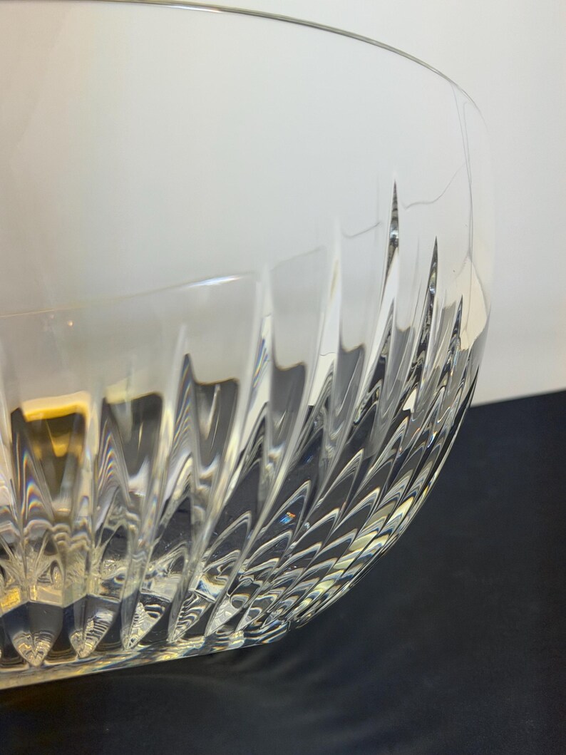 May include: A clear glass bowl with a textured, patterned design. The bowl's exterior features a series of angled, vertical cuts that create a repeating pattern. The bowl is partially lit, with light reflecting off the textured surface.