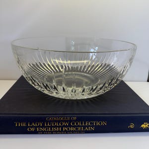 May include: A clear glass bowl with vertical ribbed detailing. The bowl is sitting on a dark blue book with gold lettering that reads "Catalogue of The Lady Ludlow Collection of English Porcelain".