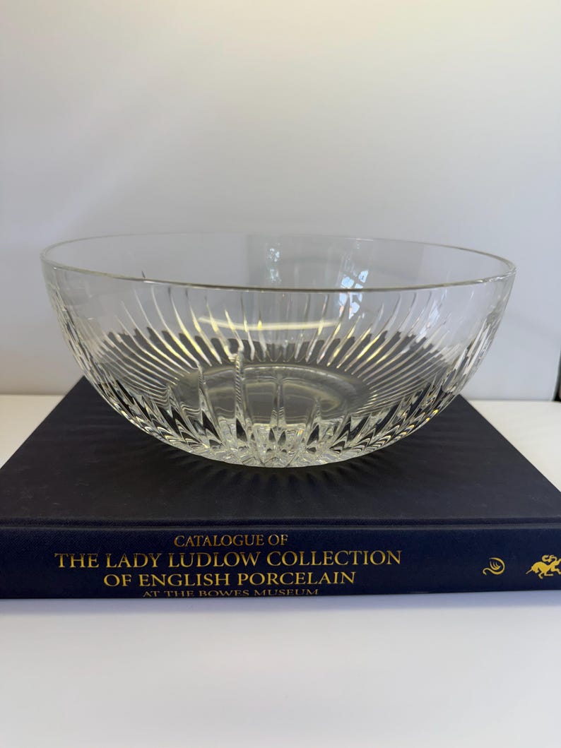 May include: A clear glass bowl with vertical ribbed detailing. The bowl is sitting on a dark blue book with gold lettering that reads "Catalogue of The Lady Ludlow Collection of English Porcelain".