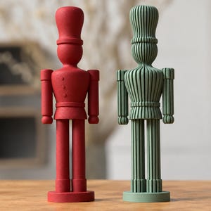 Puede incluir: Dos figuras de cascanueces de pie. La figura de la izquierda es de color rojo mate sólido con una superficie lisa. La figura de la derecha es de color verde salvia y presenta una textura distintiva de nervaduras o listones verticales.