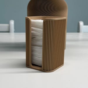 Peut inclure: Un porte-coton en bois marron à lattes verticales, avec un dessus et un dessous incurvés, rempli de tampons de coton blancs. Un vase cannelé marron assorti est en arrière-plan. Les objets sont sur une surface blanche avec un fond bleu clair.