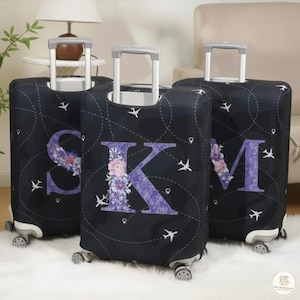 Könnte beinhalten: Drei schwarze Kofferhüllen mit den Buchstaben S, K und M in Lila, jeweils mit floralen Designs verziert. Die Hüllen haben ein Reisethemamotiv mit Flugzeugsymbolen und gepunkteten Linien und sind für Rollkoffer konzipiert.