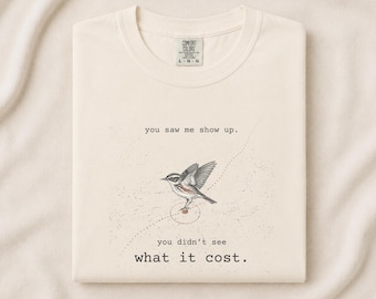 Może przedstawiać: Kremowy t-shirt z czarno-białym rysunkiem liniowym małego ptaka w locie. Tekst nad ptakiem brzmi, you saw me show up., a tekst poniżej brzmi, you didn't see what it cost. Koszulka posiada klasyczny okrągły dekolt pod szyją.