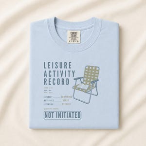 May include: A light blue t-shirt featuring a graphic of a woven lawn chair with an open book on the seat. The text reads LEISURE ACTIVITY RECORD, FORM L-17, REC. NO: RIC-0317, INTEREST CONFIRMED, MATERIALS READY, INTENTION PRESENT, and NOT INITIATED.