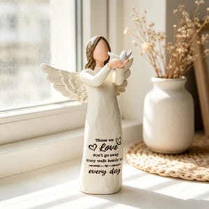 May include: A cream-colored angel figurine holding a white dove. The angel has detailed wings and brown hair. Text on the front reads "Those we love don't go away they walk beside us every day."